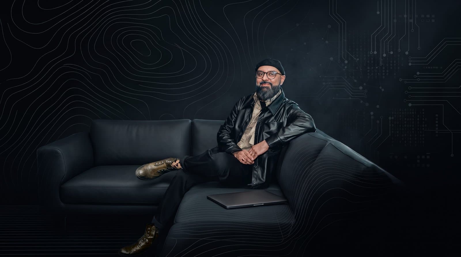 Mansur Mirani seated on a dark leather sofa in a black leather jacket, glasses and cap